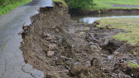 Hochwasserschaden Das Bild zeigt eine Straße, bei der der rechte Rand durch Hochwasser weggespült wurde. Im Hintergrund ist der Bach, der wieder ruhig fließt.