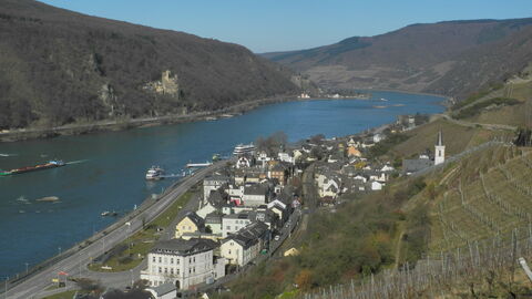 Auf dem Bild sieht man den Rhein mit seinen steilen Ufern. Zu erkennen sind Weinberge, eine Burg, eine Ortschaft und eine Straße direkt am Wasser. Schiffe fahren in der Mitte des Rheins.