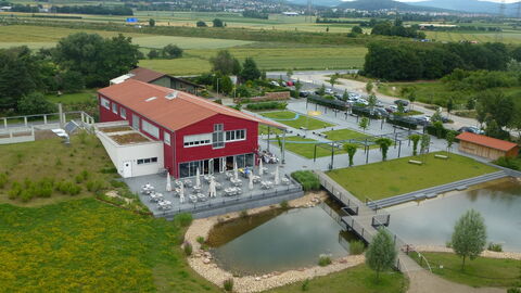 Auf dem Bild ist das besucjerzentrum des Rgionalparks RheinMain zu sehen. Ein rotes Gebäude mit rotem Dach, kleinere Gebäude nebendran, eine Terrasse mit vielen weißen Tischen, Stühlen und Sonnenschirmen vor dem Gebäude. Neben der terrasse ist ein Teich mit einer Brücke. Grüne Wiesen, Bäume und Hügel sind zu sehen.  