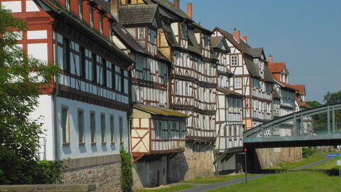 Au f dem Bild sieht man eine Häuserfront, die aus alten krummen Fachwerkhäusern besteht. Unterhalb verläuft ein Fußweg. Eine Brücke führt nach rechts.. 