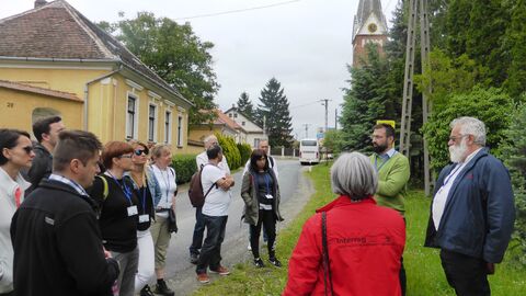 Personengruppe auf Weg, im Hintergrund Himmel, ein gelbes Haus mit Bäumen