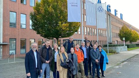 Personengruppe vor rotem Backsteingebäude, Baum und Fahnenstangen mit weißen Fahnen