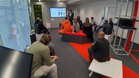 Besucher mit Blick auf Monitor und Referenten in einem Raum mit leuchtendrotem Läufer und Sitzblöcken